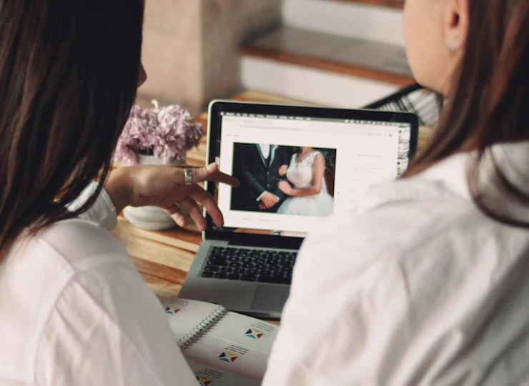 Two people in white shirts looking at wedding photo on laptop screen, with lilac flowers nearby on wooden table